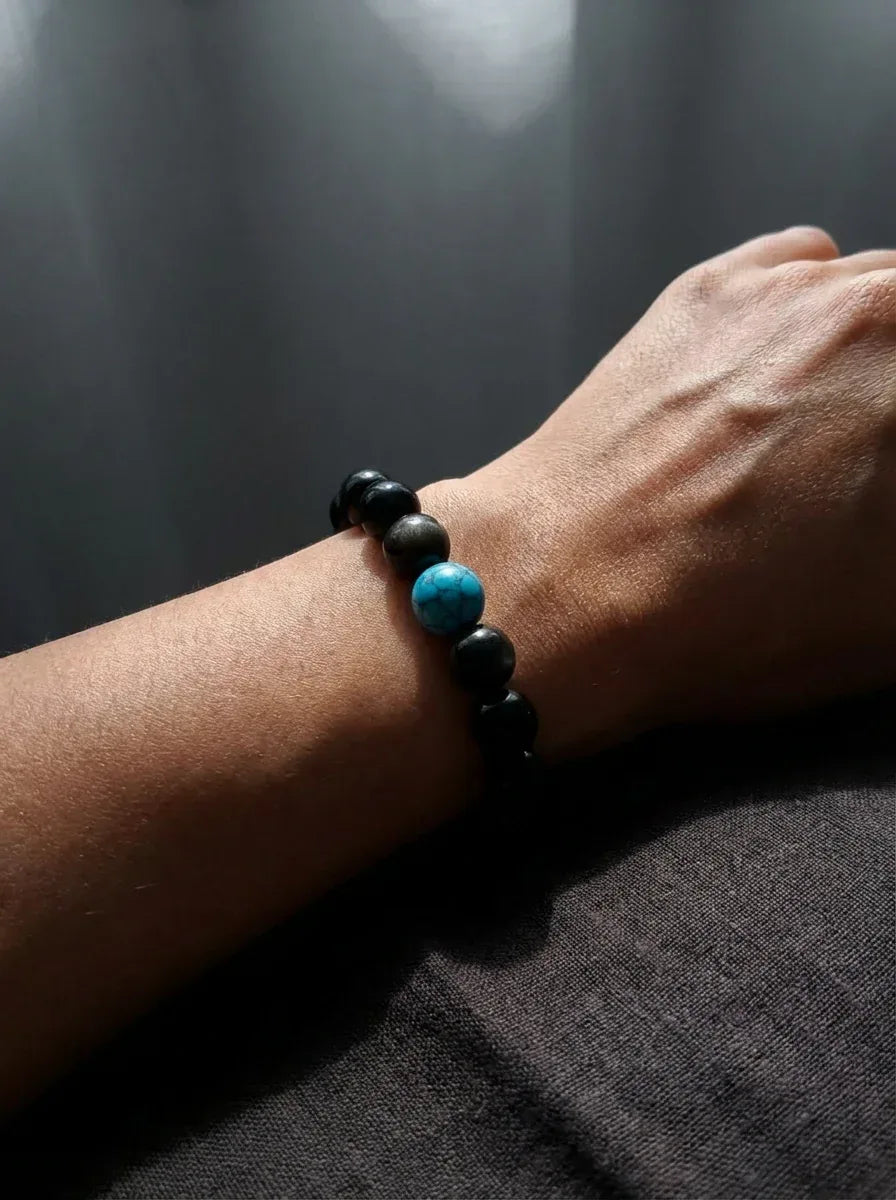 OresGreen Focus Bracelet: A minimalist black obsidian stretch bracelet with a vibrant blue turquoise focal bead worn on a man's wrist, styled under elegant side lighting for a modern zen look.