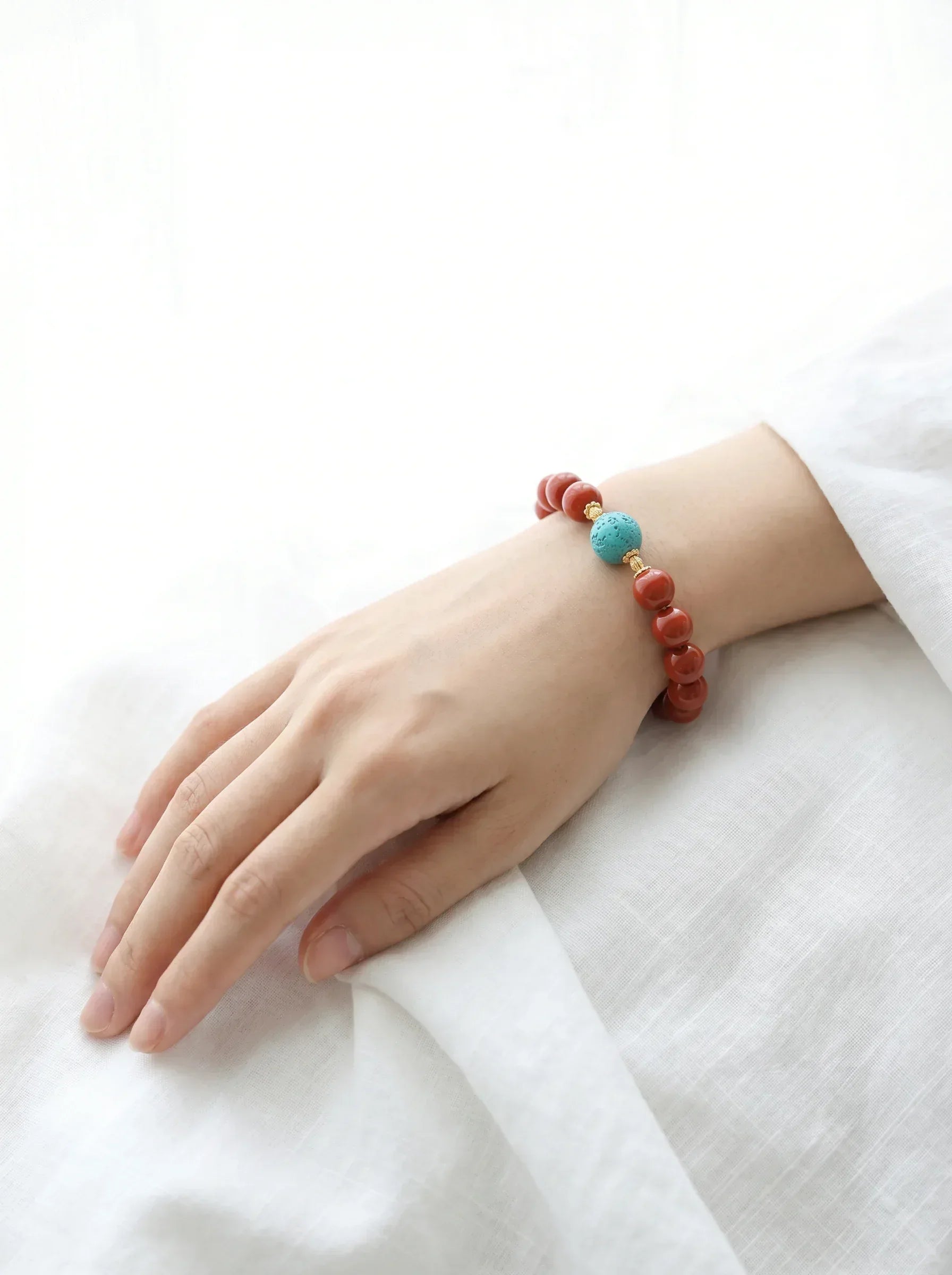 OresGreen Ember Bracelet: A minimalist red agate bead bracelet with a blue turquoise center stone and gold accents worn on a woman's wrist, styled against clean white linen clothing for a pure zen aesthetic.