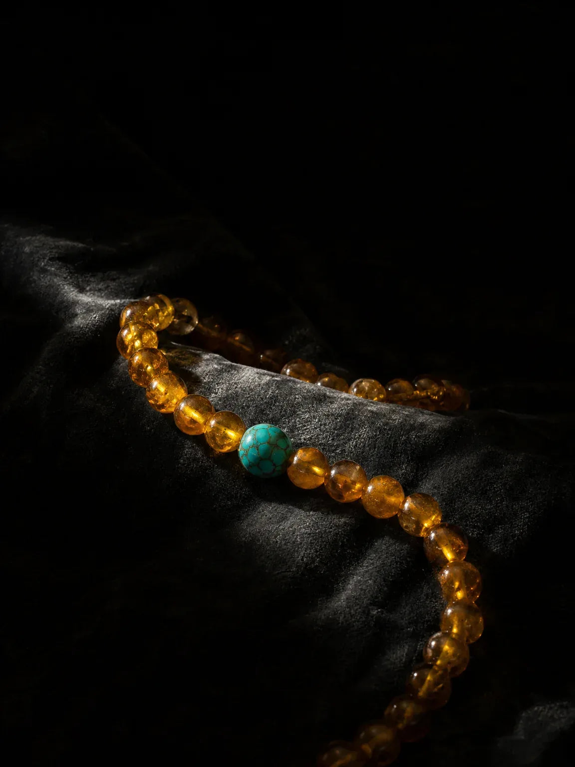 OresGreen Glow Bracelet: A vertical shot of the premium honey amber bead bracelet with a blue turquoise accent stone, beautifully glowing under a single beam of light on textured black velvet.