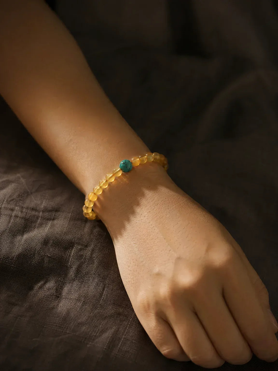 OresGreen Glow Bracelet: A minimalist honey amber bead bracelet with a single blue turquoise focal stone worn on a woman's wrist, styled with dark linen fabric for a serene zen look.