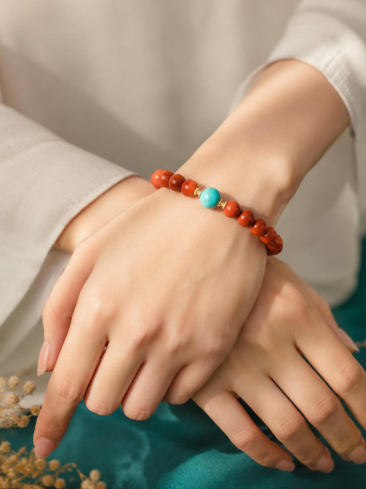 OresGreen Ember Bracelet: A minimalist red agate bead bracelet with a blue turquoise center stone and gold spacers worn on a woman's wrist, styled with elegant white silk clothing.