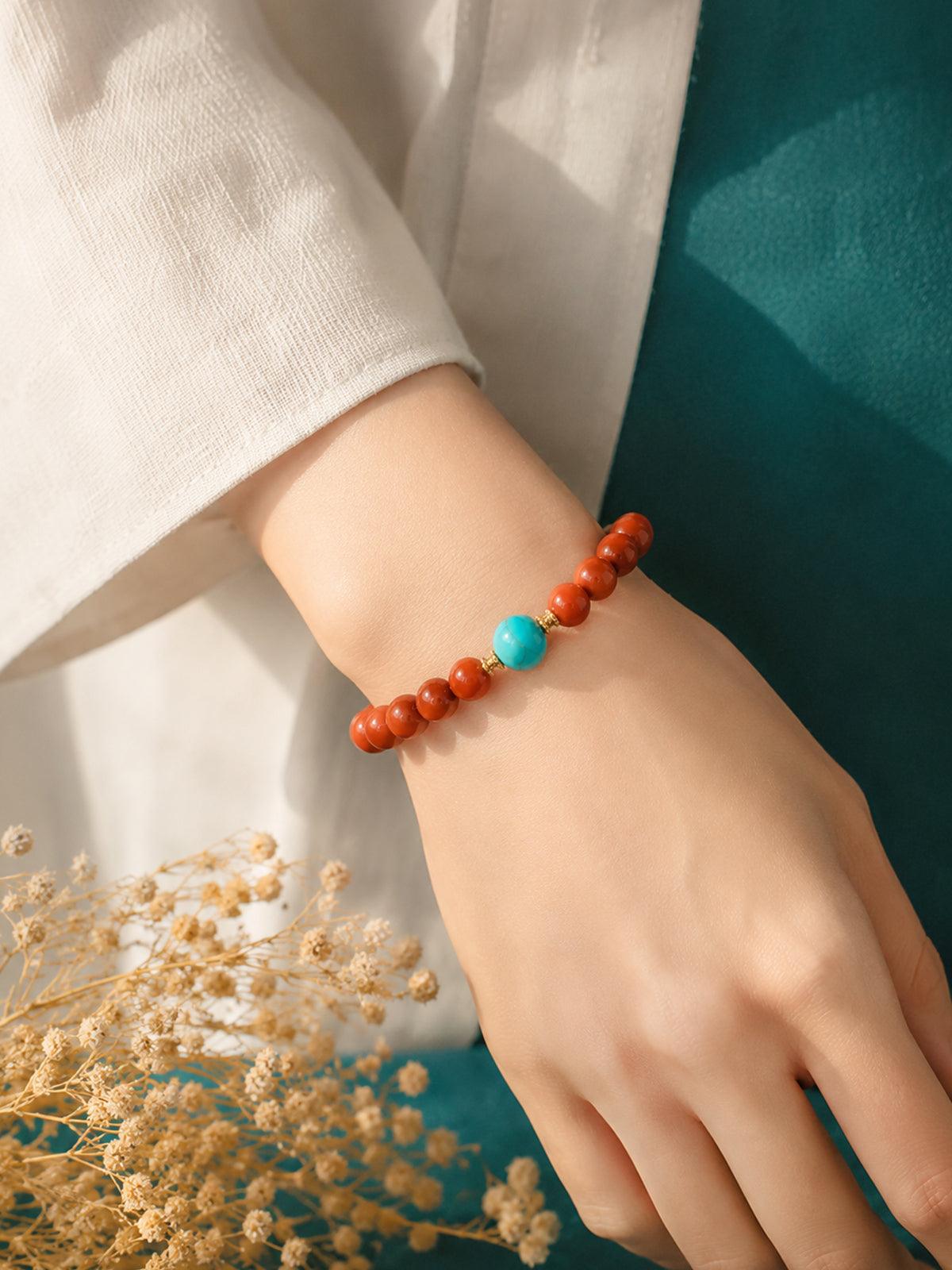 OresGreen Ember Bracelet: A minimalist red agate bead bracelet with a blue turquoise center stone and gold spacers worn on a woman's wrist, styled with elegant white silk clothing.