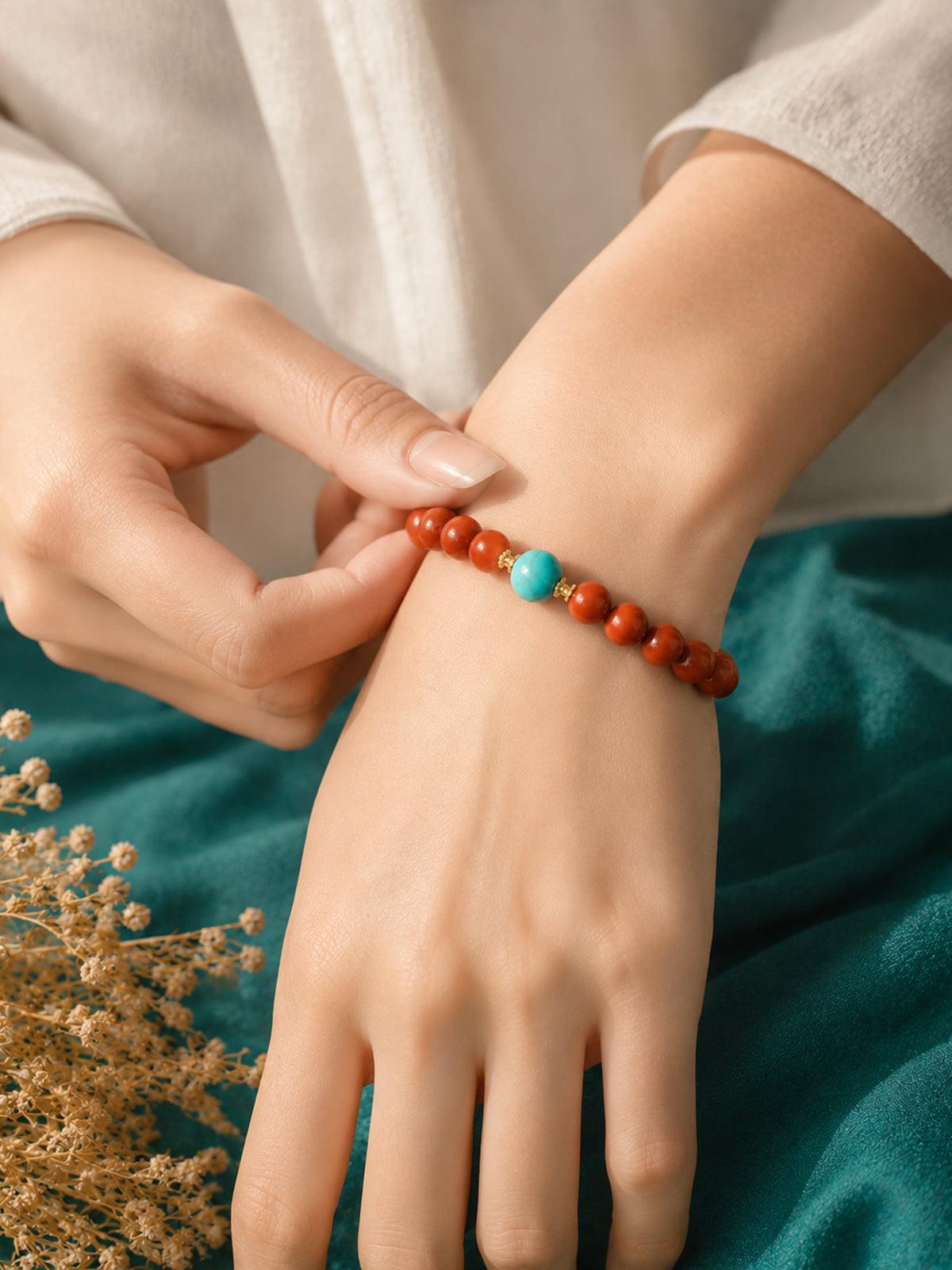 OresGreen Ember Bracelet: A minimalist red agate bead bracelet with a blue turquoise center stone and gold spacers worn on a woman's wrist, styled with elegant white silk clothing.