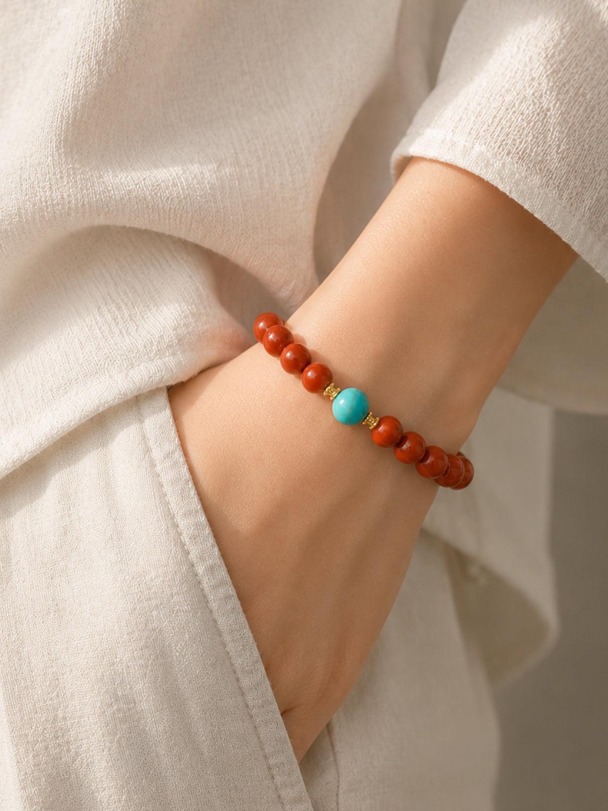 OresGreen Ember Bracelet: A minimalist red agate bead bracelet with a blue turquoise center stone and gold spacers worn on a woman's wrist, styled with elegant white silk clothing.