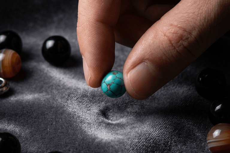OresGreen handcrafted craftsmanship: A macro close-up shot of a artisan's fingers holding a genuine blue turquoise bead with natural brown spiderweb matrix, displayed over a premium grey velvet cloth.